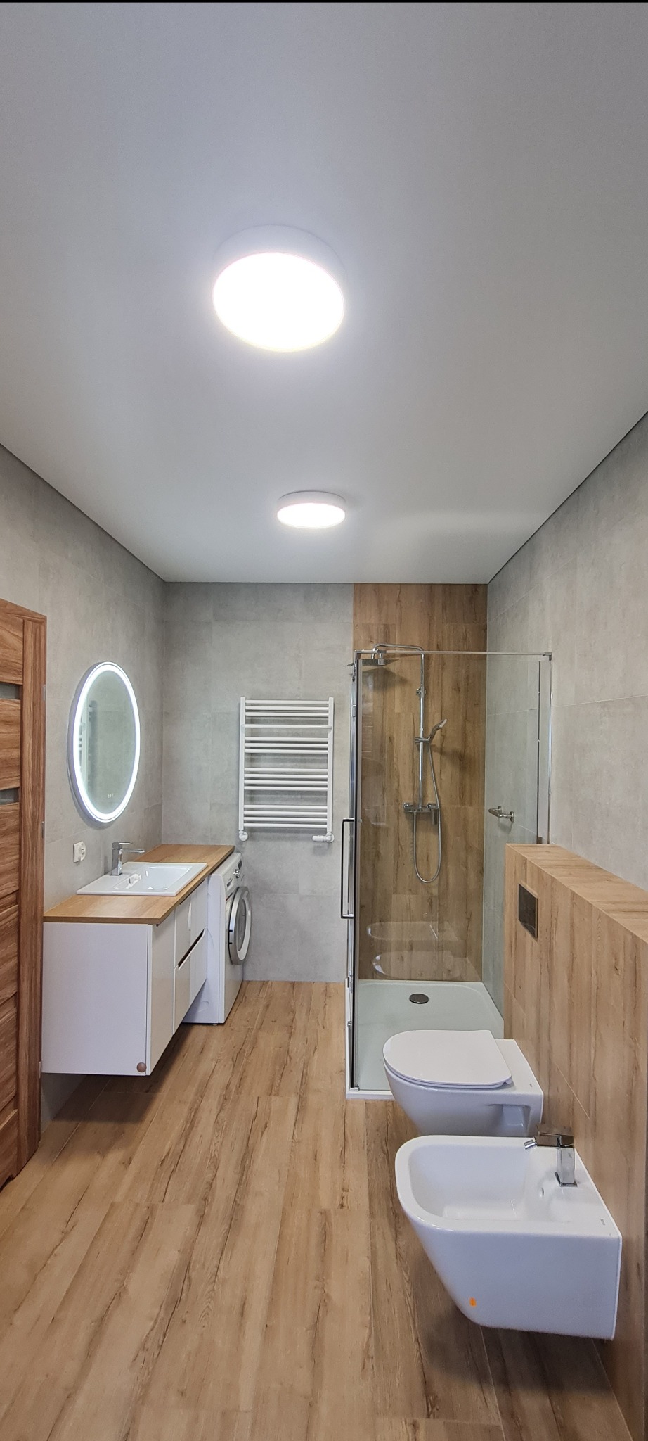 Nowoczesna łazienka z kabiną prysznicową, umywalką z szafką, toaletą i bidetem. Jasne płytki na ścianach i drewnopodobna podłoga. Okrągłe lampy sufitowe.