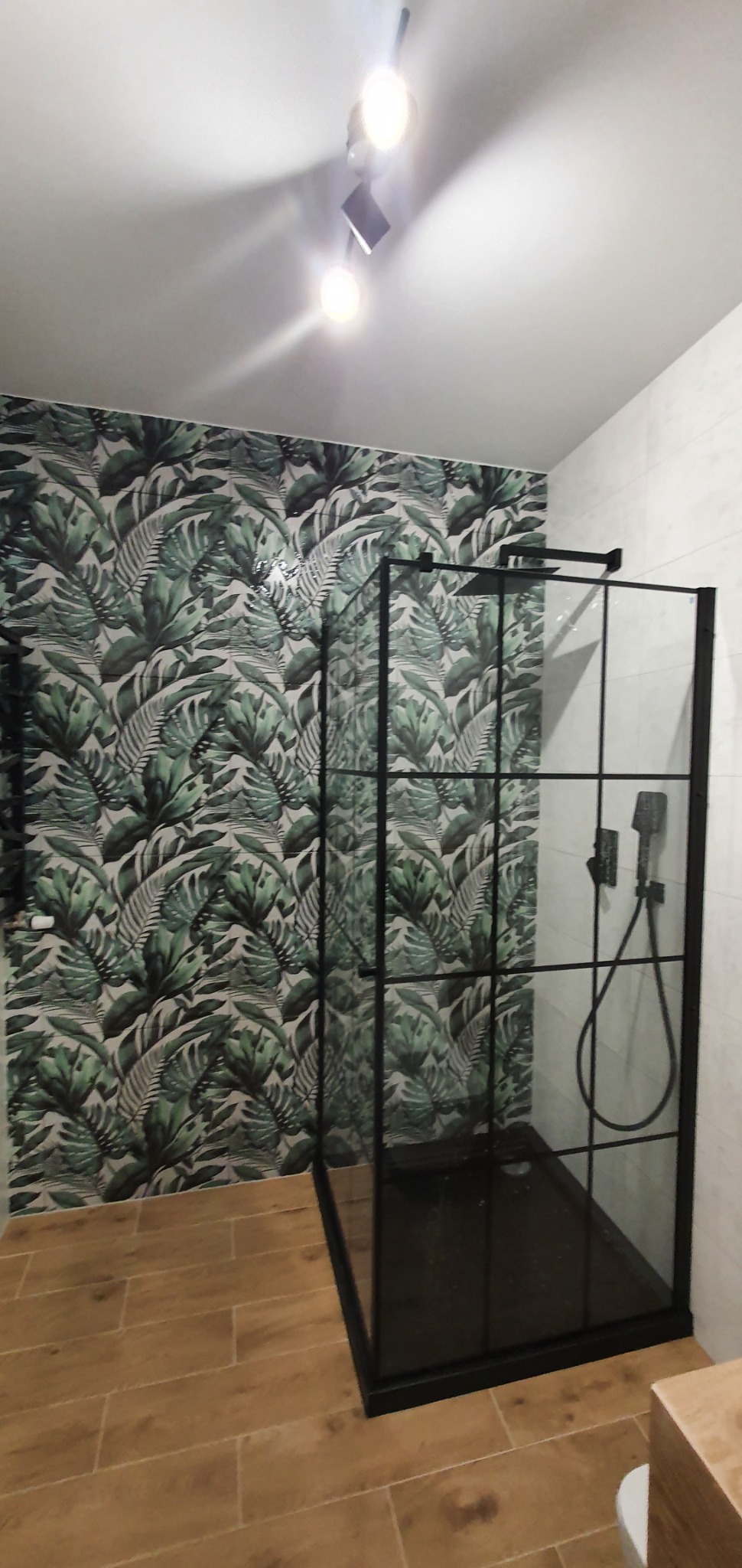 Nowoczesna kabina prysznicowa typu walk-in z czarnymi profilami na tle ściany wyłożonej płytkami z motywem tropikalnych liści, drewnopodobne płytki podłogowe i punktowe oświetlenie sufitowe.