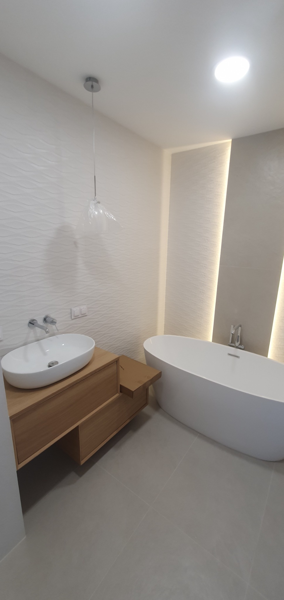 Nowoczesna łazienka z wolnostojącą wanną i umywalką nablatową na drewnianej szafce, oświetlenie punktowe i dekoracyjna ściana z subtelnym wzorem fal.