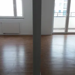 Puste, jasne pomieszczenie z parkietem, grzejnikiem i balkonem widoczne przez rozsuwane drzwi balkonowe, oddzielone centralnie umieszczonym słupem.