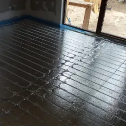 Błyszcząca folia izolacyjna z ułożonymi rurami ogrzewania podłogowego na podłożu w surowym pomieszczeniu, widok na okno z pianką uszczelniającą.