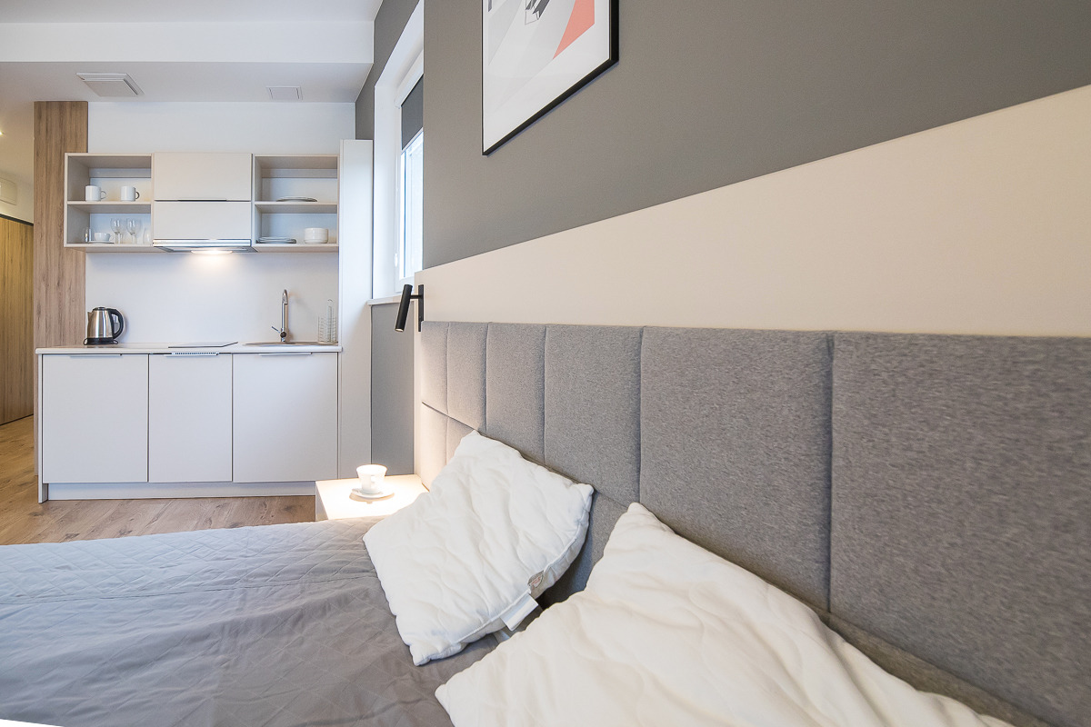 Nowoczesne studio z aneksem kuchennym, szarym wezgłowiem łóżka i minimalistycznym wystrojem, z widocznym czajnikiem elektrycznym i filiżanką na stoliku nocnym.