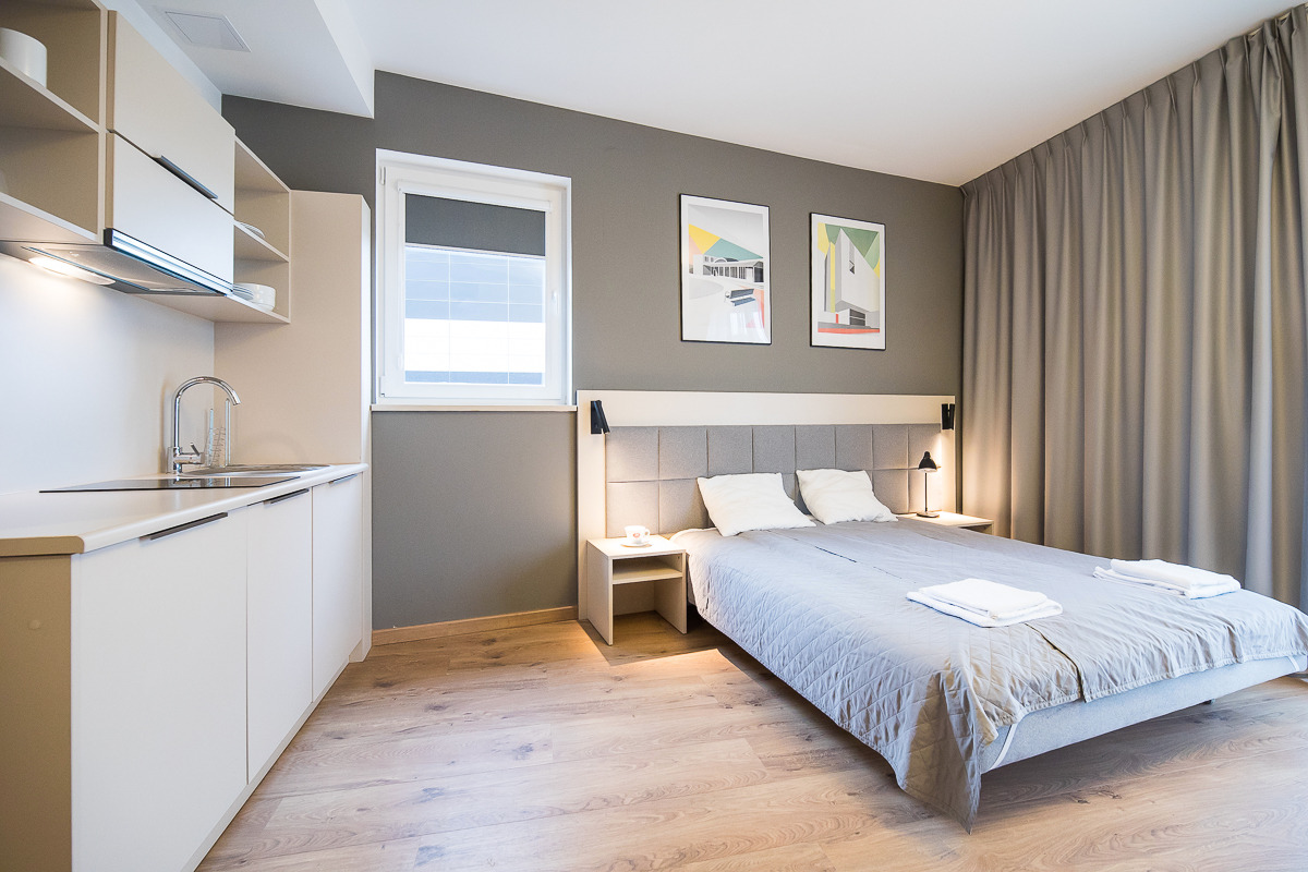 Jasne wnętrze pokoju hotelowego z aneksem kuchennym, szarymi ścianami, drewnianą podłogą, łóżkiem z szarą narzutą i białymi poduszkami, dwoma obrazami na ścianie i zasłoniętym oknem.