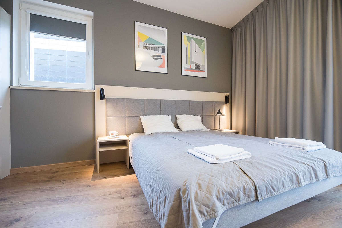 Jasnoszara sypialnia w Warszawie z podwójnym łóżkiem, szarą tapicerowaną ścianą, dwoma stolikami nocnymi z lampkami i filiżanką, oraz dwoma obrazami abstrakcyjnymi na ścianie.