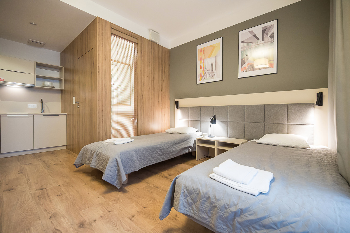 Pokój hotelowy z dwoma pojedynczymi łóżkami, aneksem kuchennym, drewnianą boazerią i abstrakcyjnymi obrazami na ścianie.