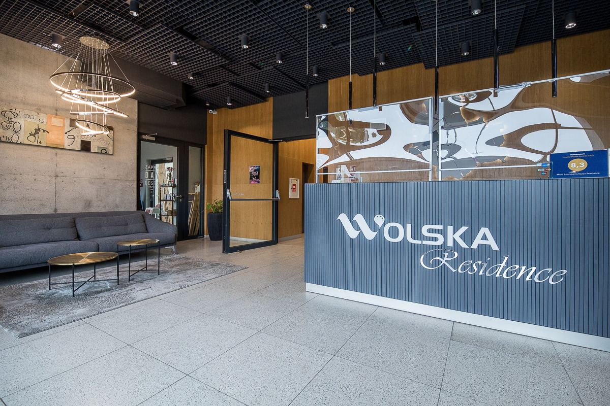 Nowoczesne wnętrze recepcji z szarą sofą, stolikami kawowymi i betonową ścianą z abstrakcyjnym obrazem, widok na wejście do budynku z przeszklonymi drzwiami.