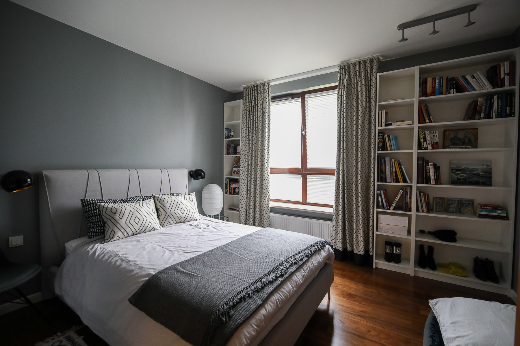 Szara sypialnia z drewnianą podłogą, łóżkiem z pikowanym zagłówkiem i geometrycznymi poduszkami, białe regały z książkami po bokach okna z żaluzjami.