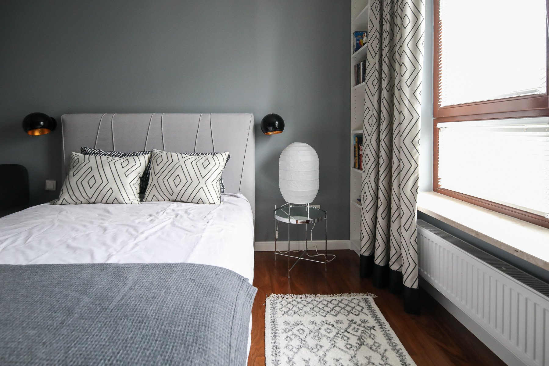 Nowoczesna sypialnia z szarą ścianą, łóżkiem z tapicerowanym zagłówkiem, geometrycznymi poduszkami, lampką nocną na metalowym stoliku, wzorzystymi zasłonami i dywanikiem.