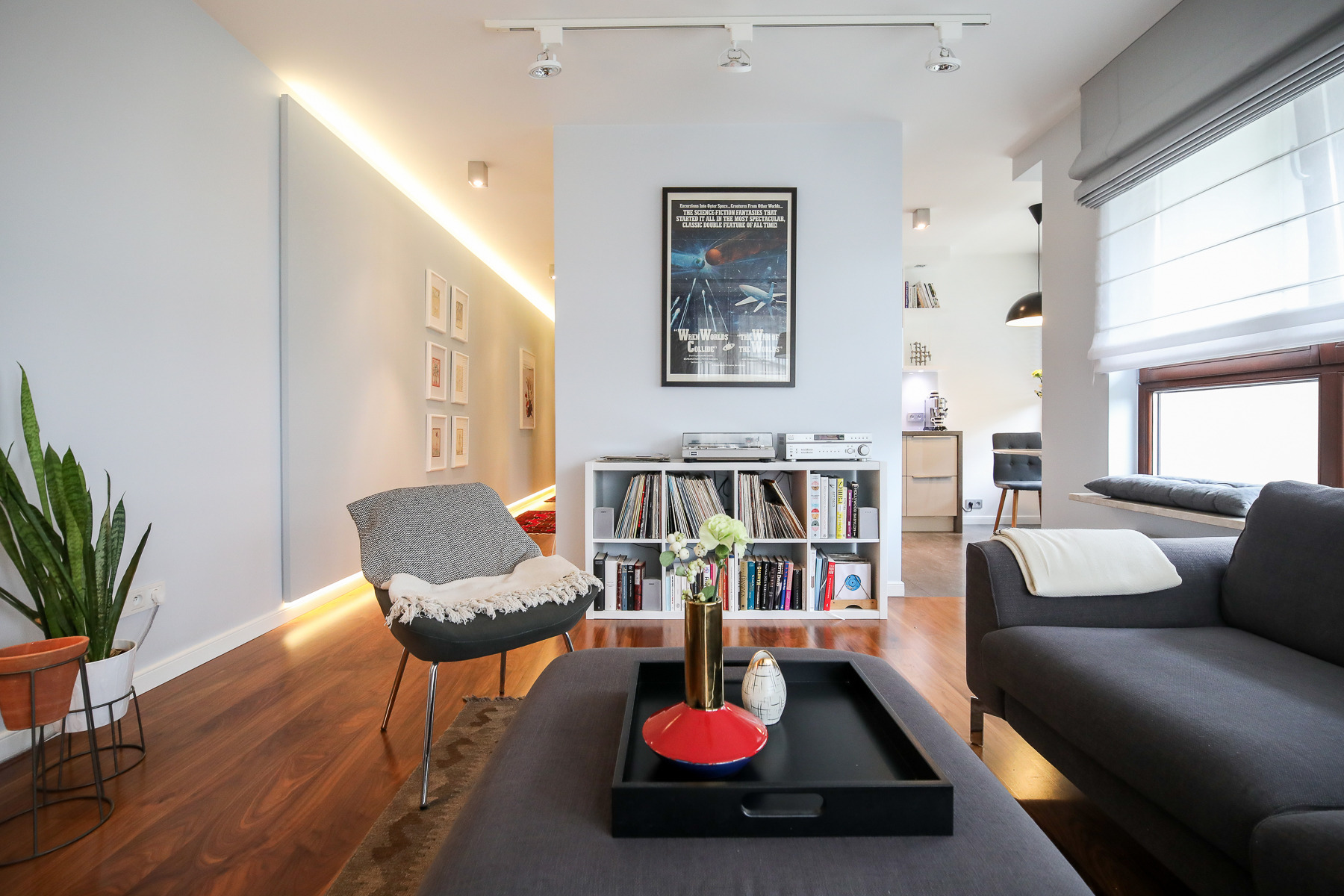 Jasne, przestronne wnętrze salonu z drewnianą podłogą, szarą sofą przy oknie z roletami rzymskimi, fotel z poduszką, biały regał z płytami i książkami oraz stolik kawowy z dekoracyjnym wazonem i tacą.