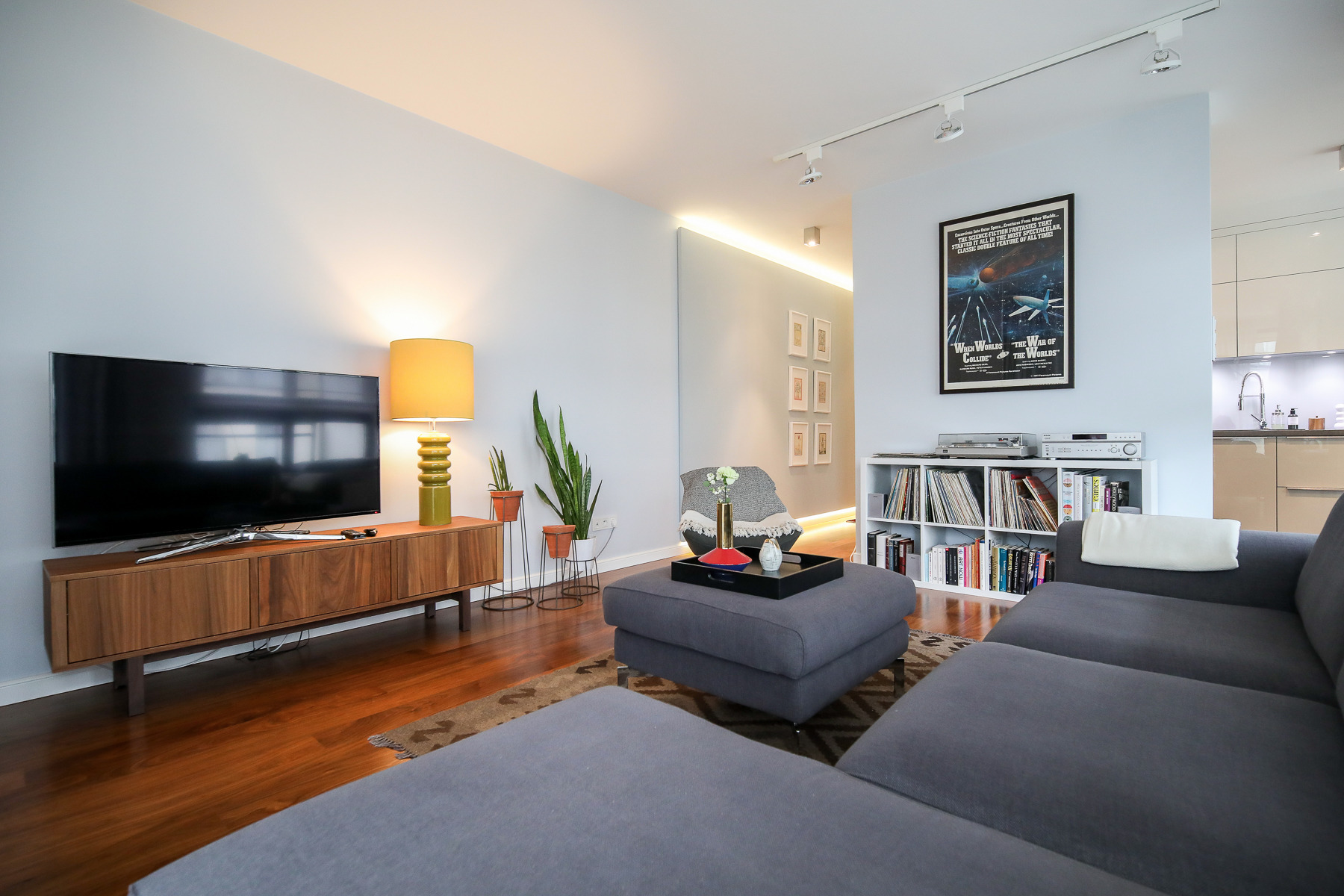 Nowoczesny salon z szarą sofą modułową, drewnianą szafką RTV z telewizorem, zieloną lampą ceramiczną i regałem na płyty winylowe z plakatem filmu science fiction na ścianie.