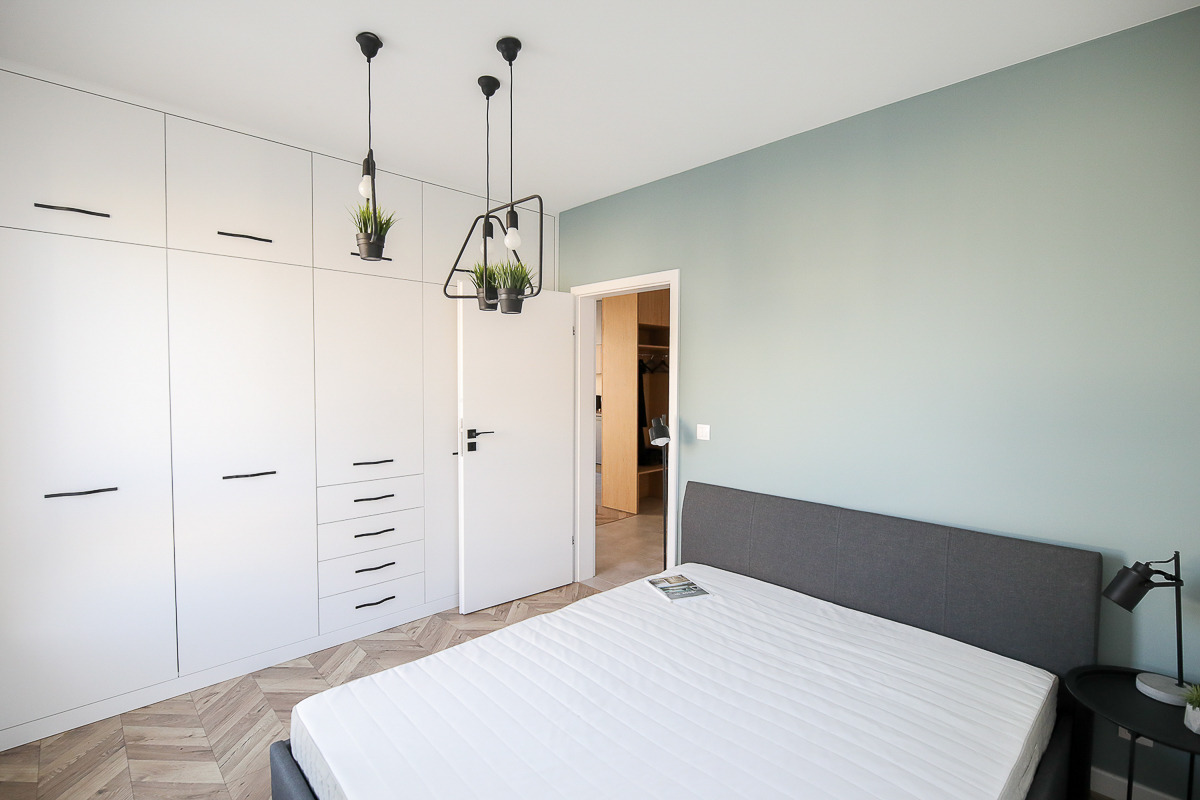 Nowoczesna sypialnia z białymi szafami w zabudowie, parkietem w jodełkę i minimalistycznymi lampami z roślinami, widok na otwarte drzwi do garderoby.