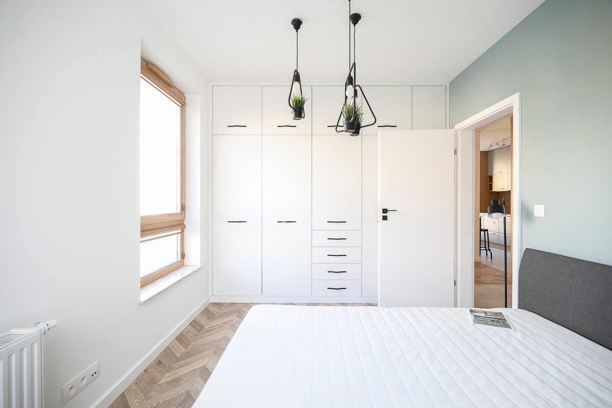 Jasna sypialnia z białymi szafami w zabudowie, drewnianą podłogą w jodełkę i designerskimi lampami wiszącymi z doniczkami.