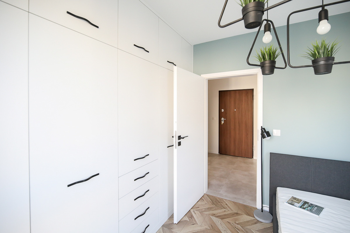 Wnętrze pokoju z białym, minimalistycznym systemem szaf z czarnymi, falistymi uchwytami, otwartymi drzwiami i lampą sufitową z doniczkami z roślinami, widok na korytarz z drewnianymi drzwiami...