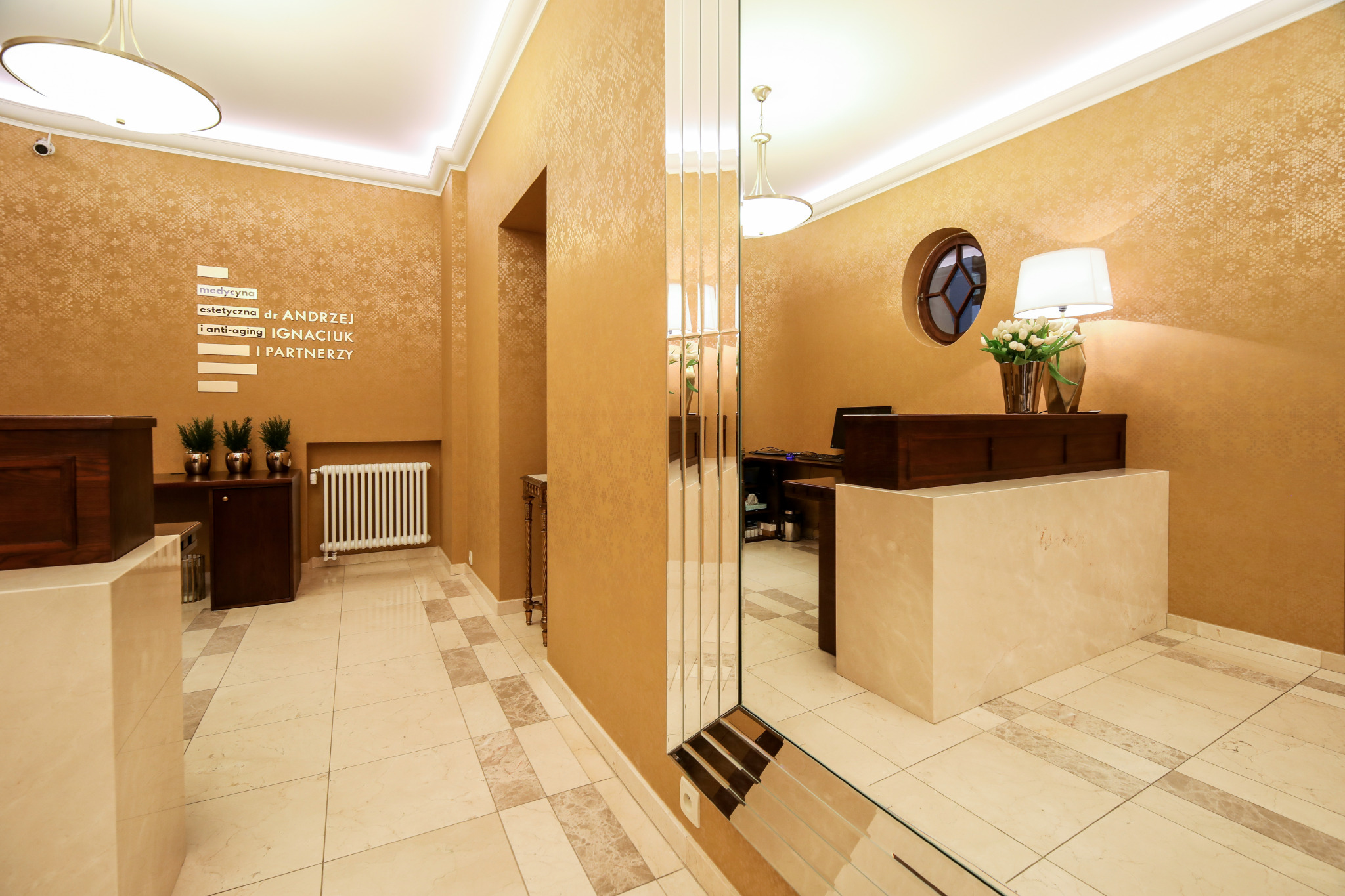 Eleganckie wnętrze recepcji z marmurową podłogą, złotymi ścianami z subtelnym wzorem, drewnianą ladą, lustrem w wąskich pasach i dekoracyjnym oświetleniem.