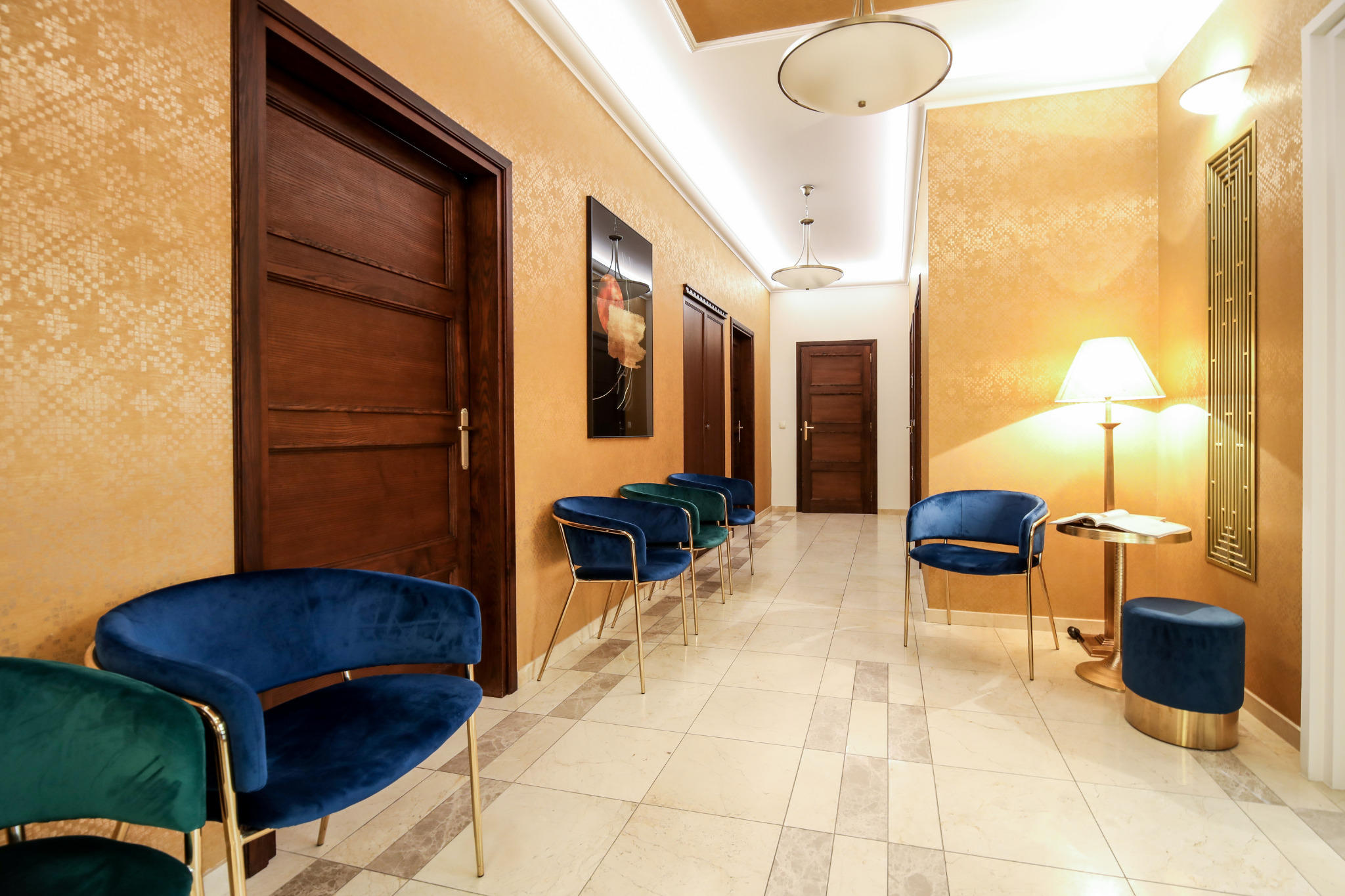 Elegancka poczekalnia z krzesłami obitymi aksamitem w odcieniach granatu i zieleni, złotymi nogami i detalami, oświetlona lampami w stylu art deco, z drewnianymi drzwiami i wzorzystymi tapetami...