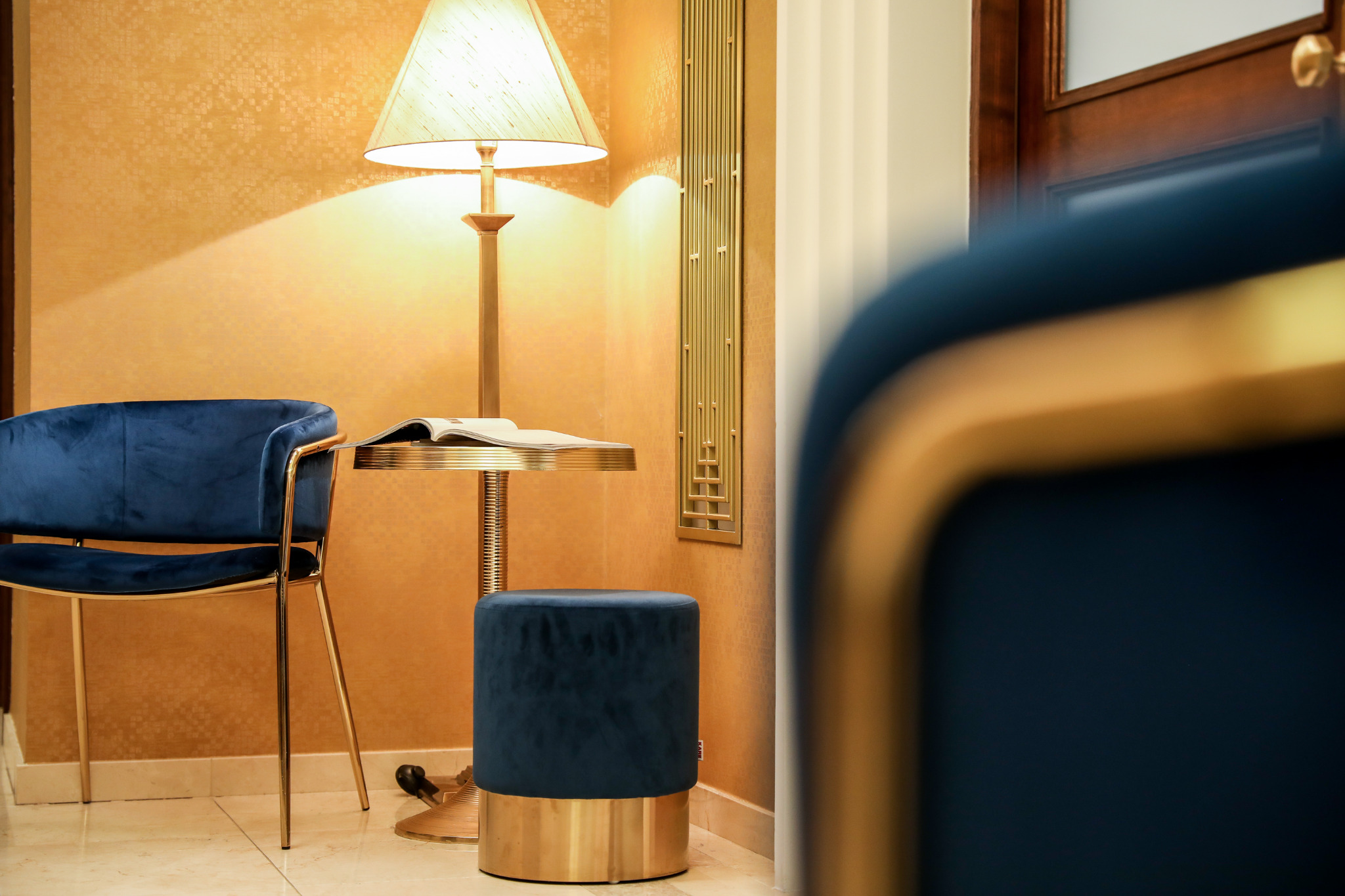 Niebieski, welurowy fotel i pufa z elementami w kolorze złotym, ustawione w pokoju z dekoracyjną lampą podłogową i ozdobnym panelem ściennym.