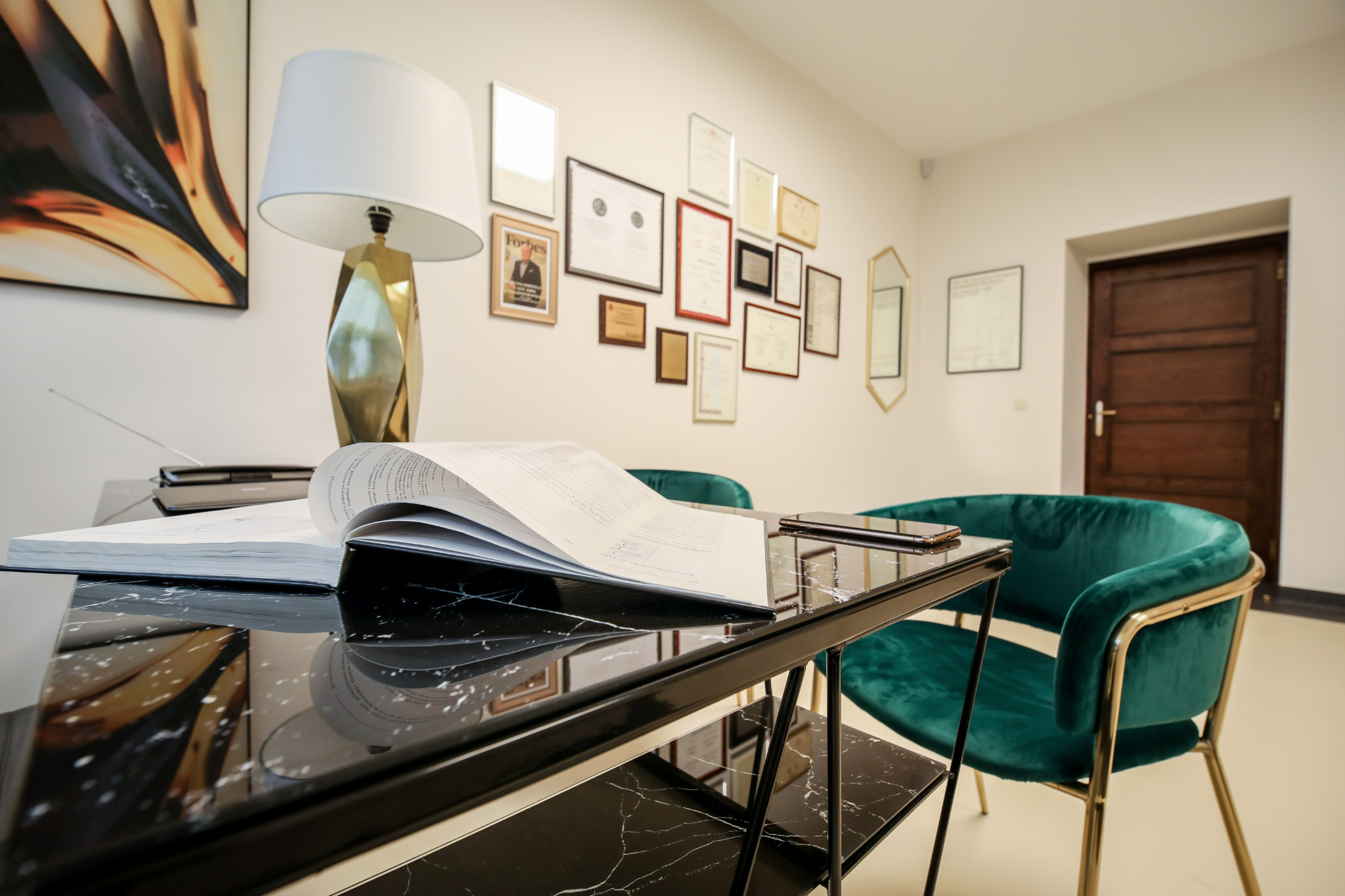 Eleganckie biuro z czarnym marmurowym biurkiem, otwartą książką, zielonym krzesłem welurowym ze złotymi nogami i ścianą z certyfikatami i nagrodami.