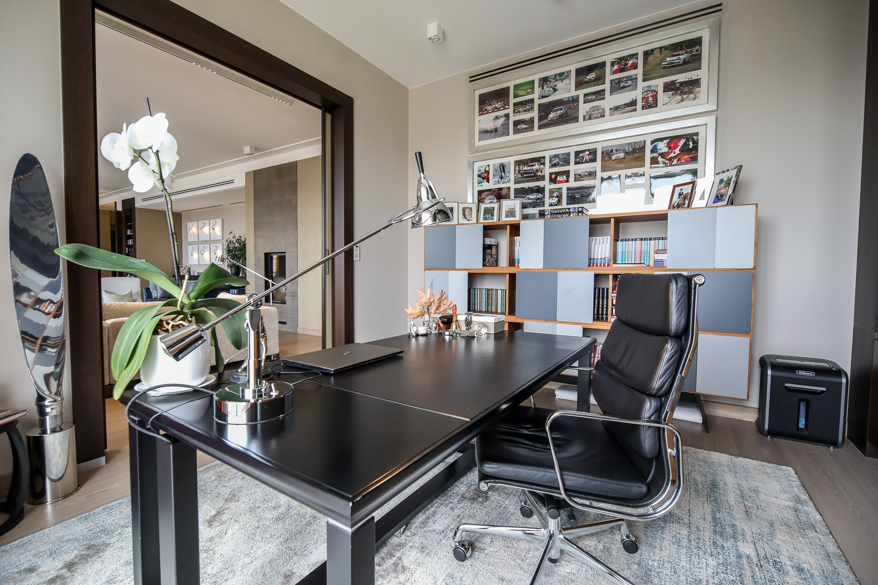 Nowoczesne biuro domowe z czarnym biurkiem, skórzanym fotelem, lampą biurkową i dekoracyjnymi elementami, takimi jak orchidea i metalowa rzeźba, na tle regału z książkami i zdjęciami wyścigowymi.