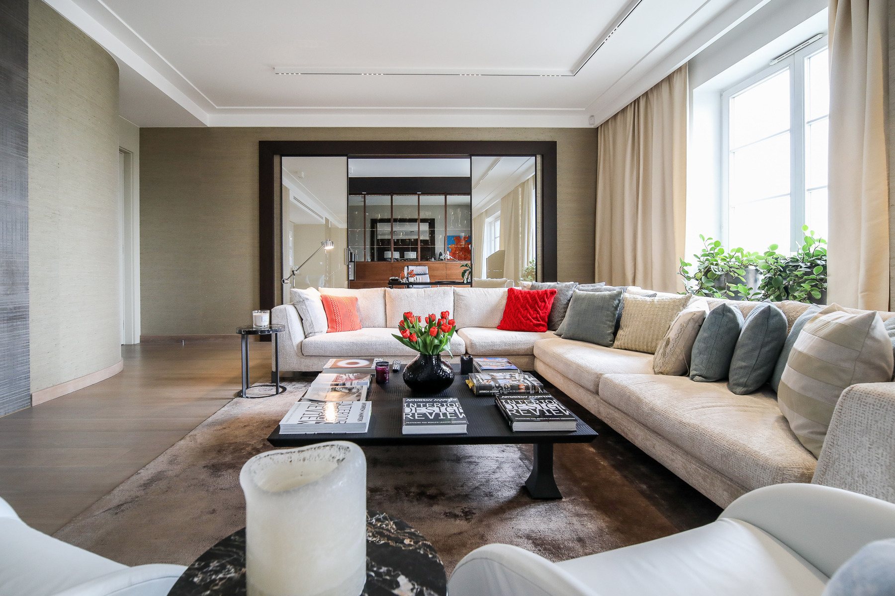 Jasny salon z narożną sofą, czarnym stolikiem kawowym z książkami i wazonem czerwonych tulipanów, duże okno z roślinami i lustro w ciemnej ramie.