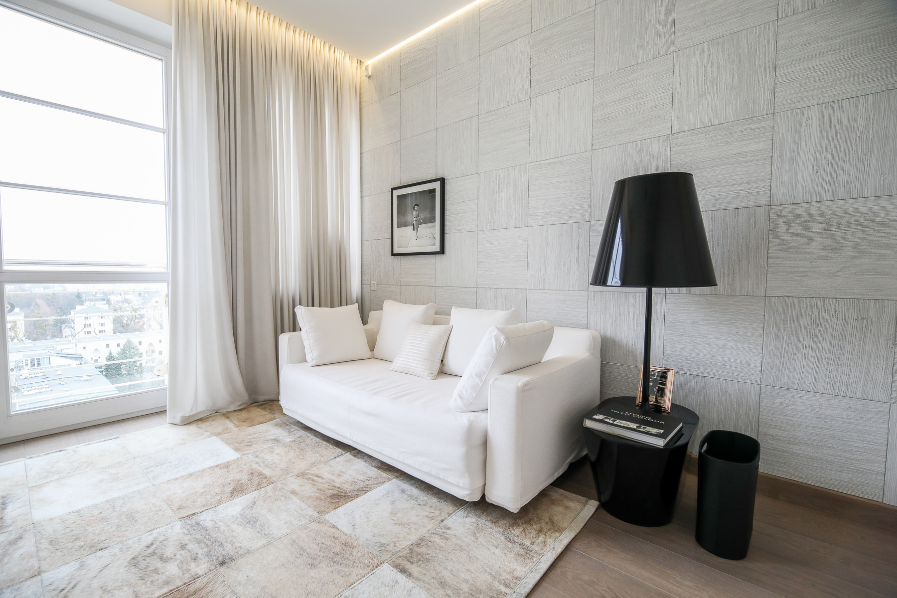 Jasne wnętrze pokoju dziennego z białym narożnikiem, poduszkami, lampą podłogową i dywanem w odcieniach beżu i brązu, widok na miasto przez duże okno.