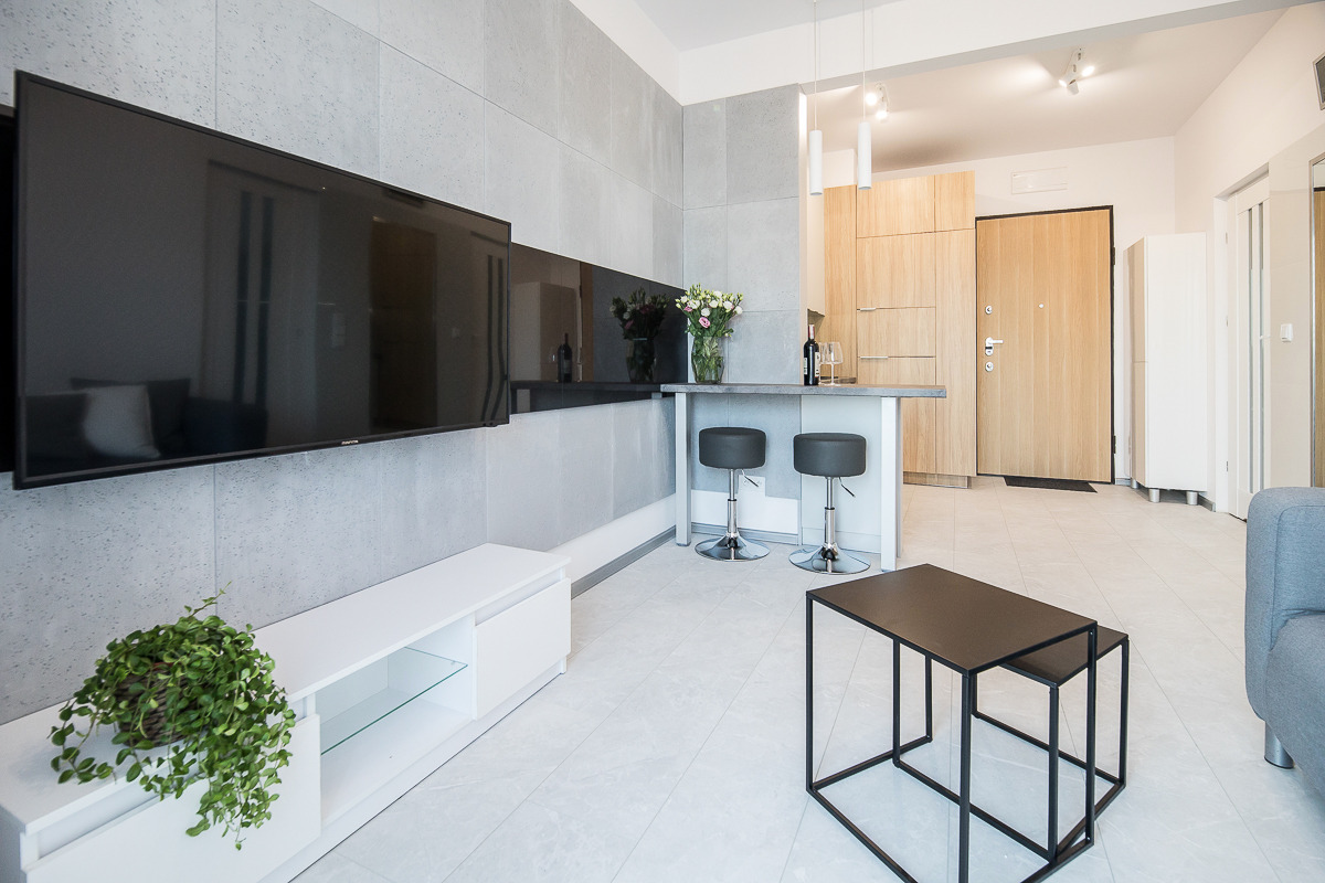 Nowoczesne wnętrze mieszkania z szarymi ścianami, dużym telewizorem, białymi meblami, barkiem z hokerami i stolikiem kawowym o minimalistycznej konstrukcji.