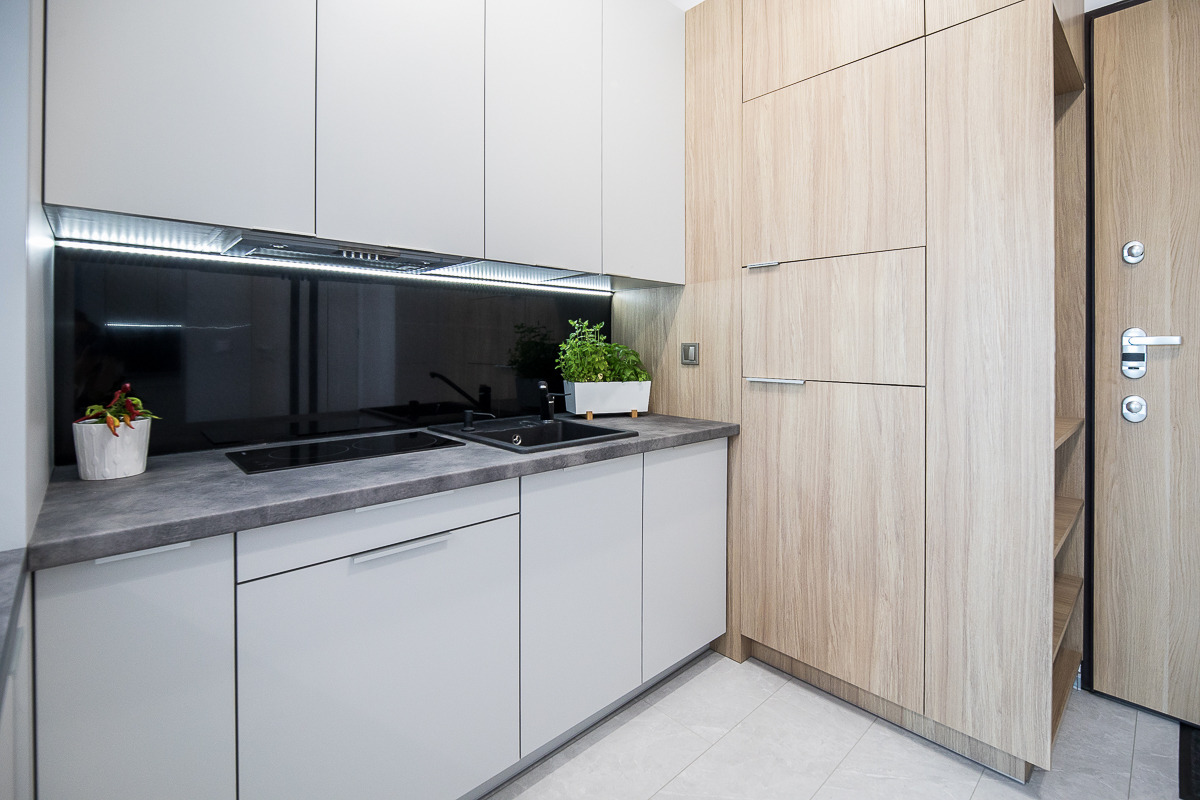 Nowoczesna kuchnia z szarymi szafkami, czarnym panelem nad blatem, drewnianymi szafkami po prawej i roślinami w doniczkach na blacie.