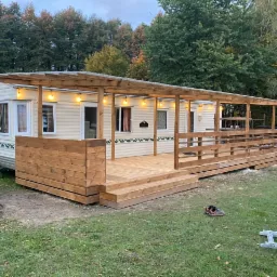 Mobilny dom z dobudowanym drewnianym tarasem i zadaszeniem, oświetlonym girlandą żar&oacute;wek, widoczne narzędzia i resztki materiał&oacute;w budowlanych.