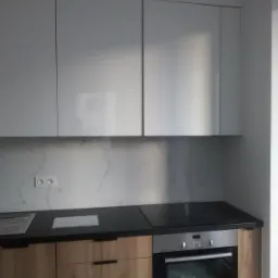 Nowoczesna zabudowa kuchenna z drewnianymi szafkami dolnymi, czarnym blatem i białymi szafkami górnymi na tle marmurowej ściany z gniazdkami elektrycznymi.