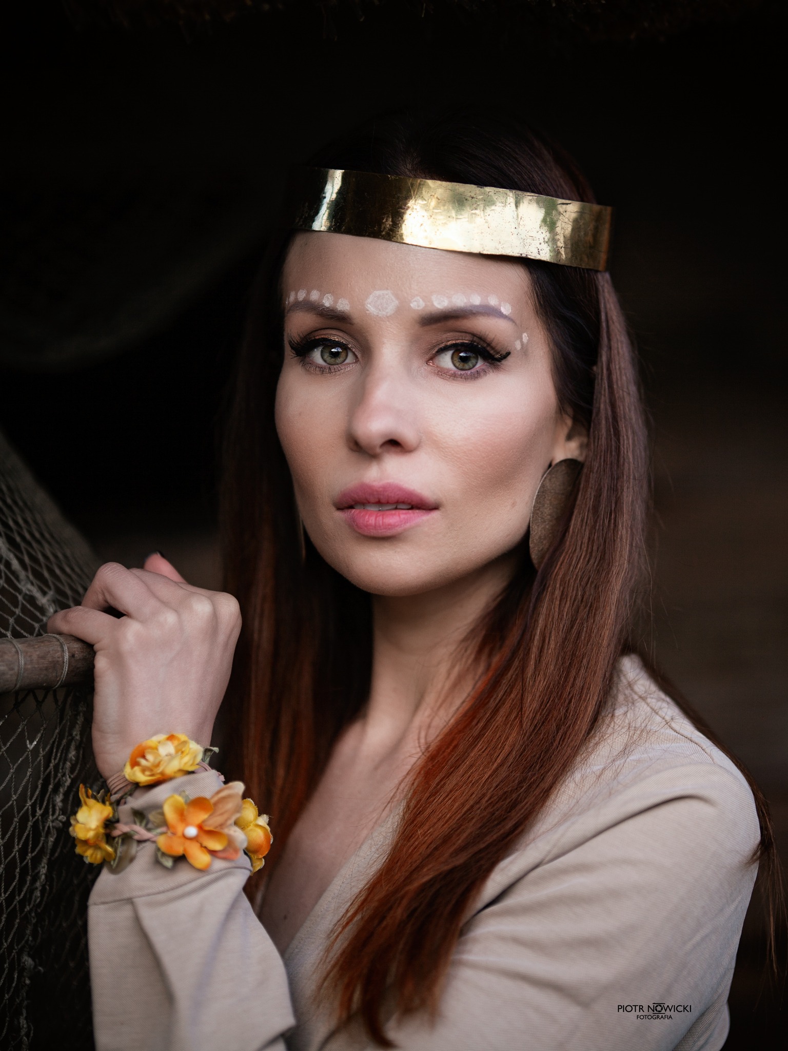 Portret kobiety z etnicznym makijażem, złotą opaską na głowie i bransoletką z kwiatów, pozującej na tle ciemnej ściany i rybackiej sieci.