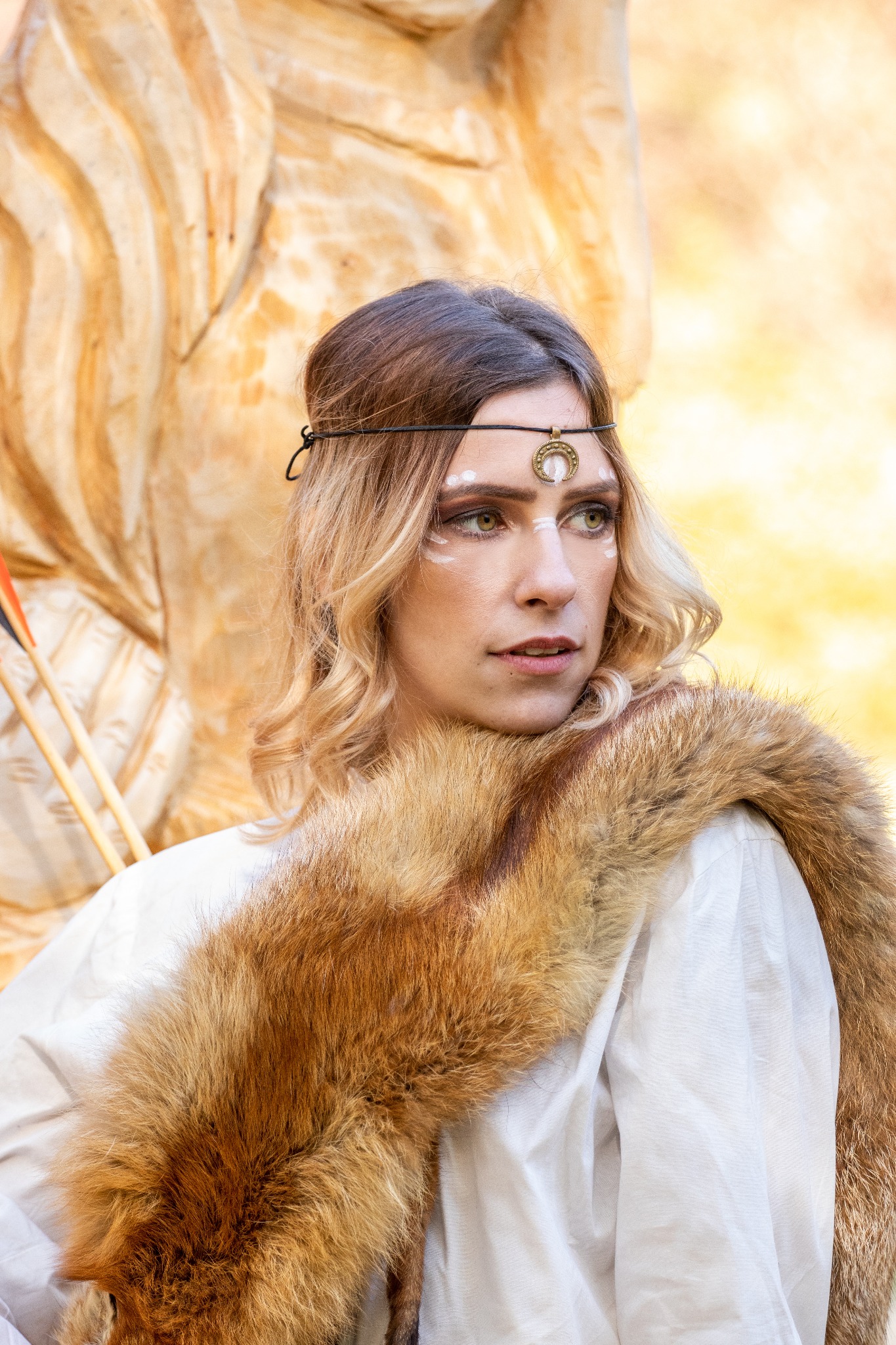 Portret kobiety z jasnymi włosami i futrzanym szalem, z delikatnym, białym malunkiem na twarzy i ozdobą na głowie w stylu etnicznym, na tle drewnianej rzeźby.