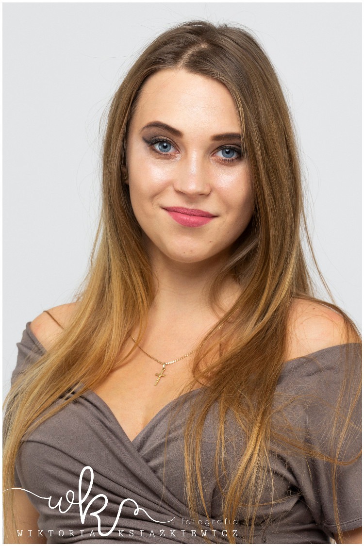 Portret kobiety z subtelnym makijażem oczu i ust, złoty łańcuszek z krzyżykiem, jasne tło.