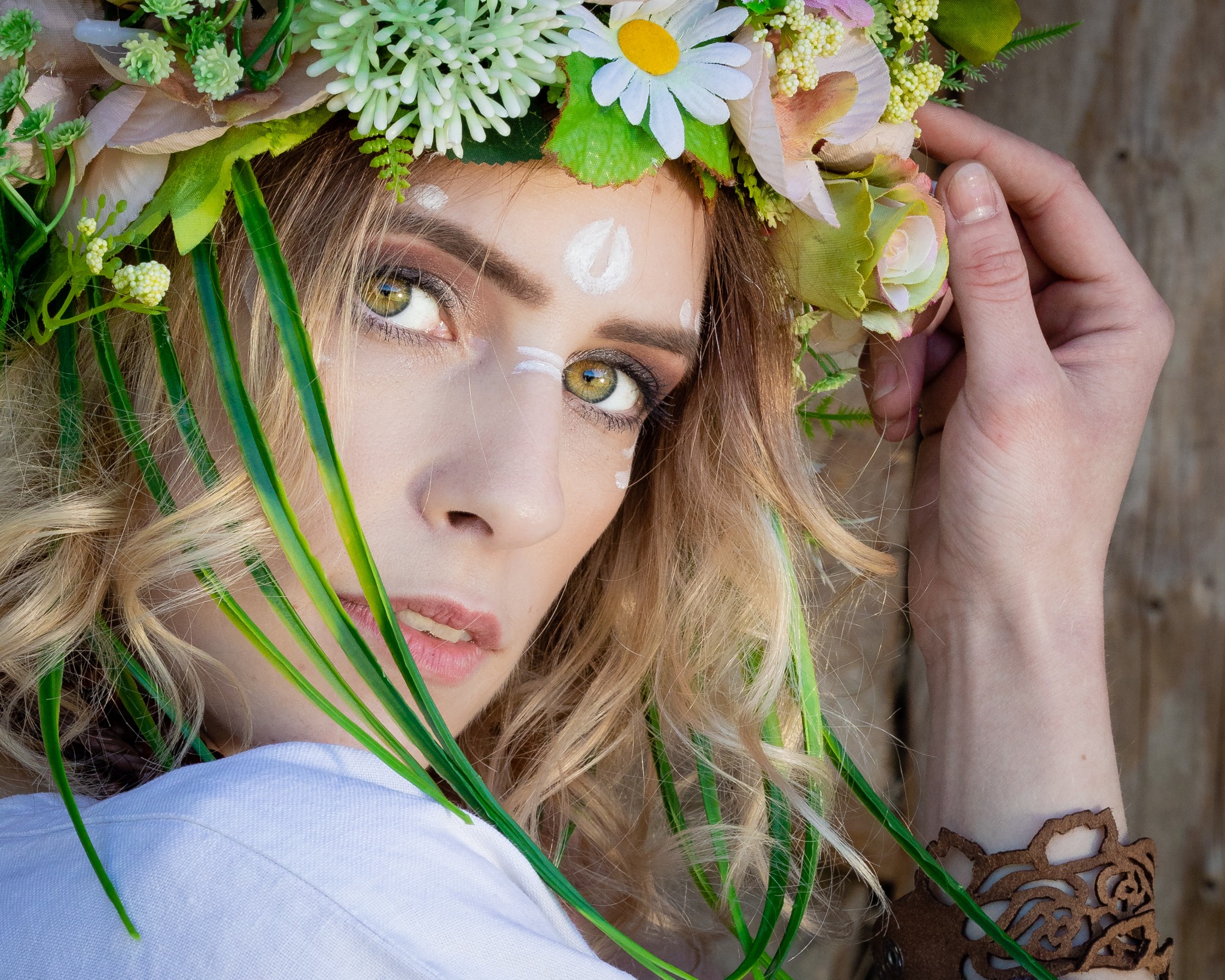 Portret kobiety z kwiatowym wiankiem, delikatnym makijażem oczu i białymi akcentami na czole, zamyślone spojrzenie w obiektyw, brązowa bransoletka na nadgarstku.