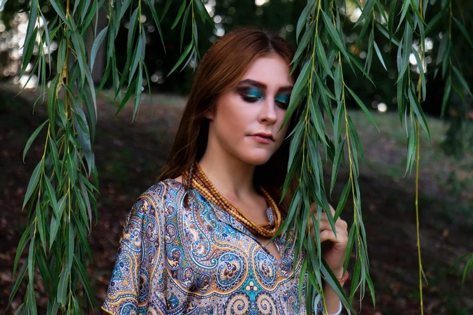 Portret kobiety z intensywnym turkusowym cieniem na powiekach, pozującej na tle zieleni i zwisających gałęzi wierzby.
