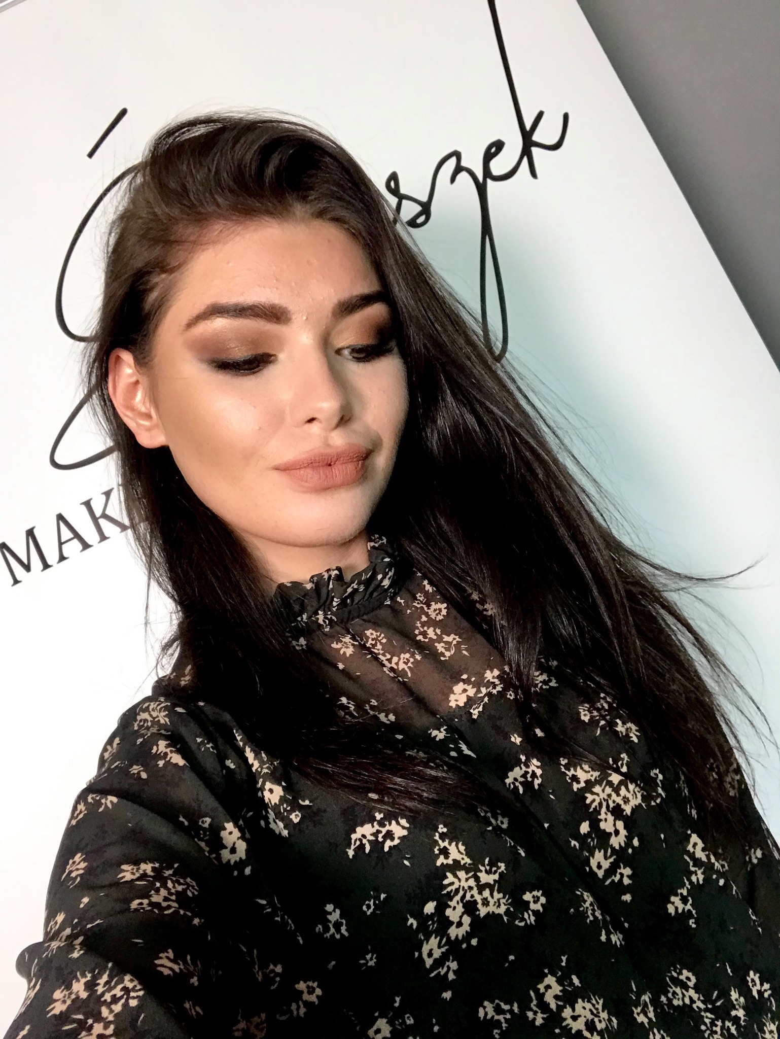 Portret kobiety z profesjonalnym makijażem oczu w odcieniach brązu i ust w kolorze nude, na tle białej ściany z napisem 'MAK' i logiem.