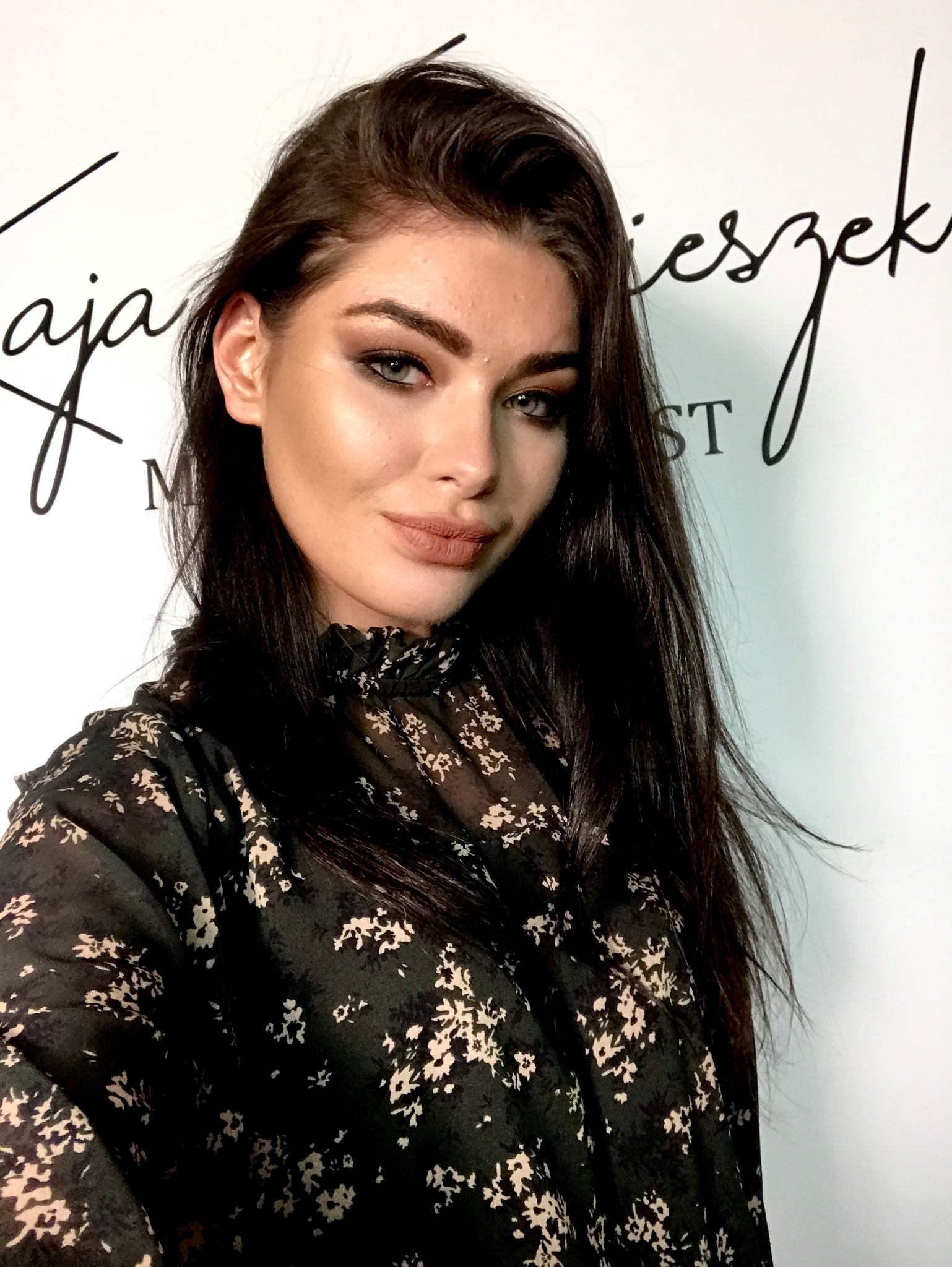 Portret kobiety z profesjonalnym, wyrazistym makijażem oczu i ust w odcieniach nude, na tle białej ściany z czarnym napisem stylizowanym na odręczny.