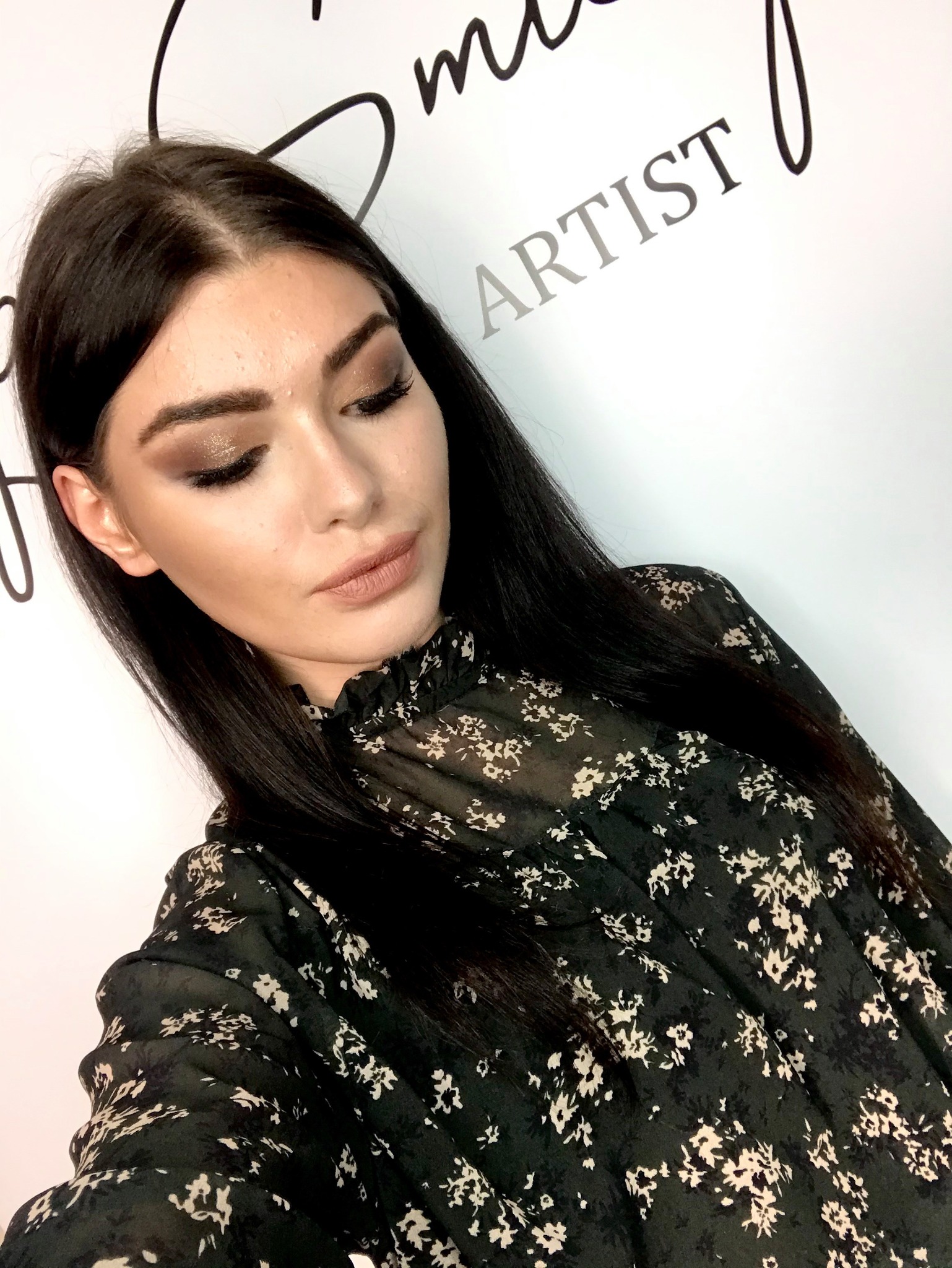 Portret kobiety z profesjonalnym makijażem oczu w odcieniach brązu i ust w kolorze nude, na tle białej ściany z napisem 'Smiling Artist'.