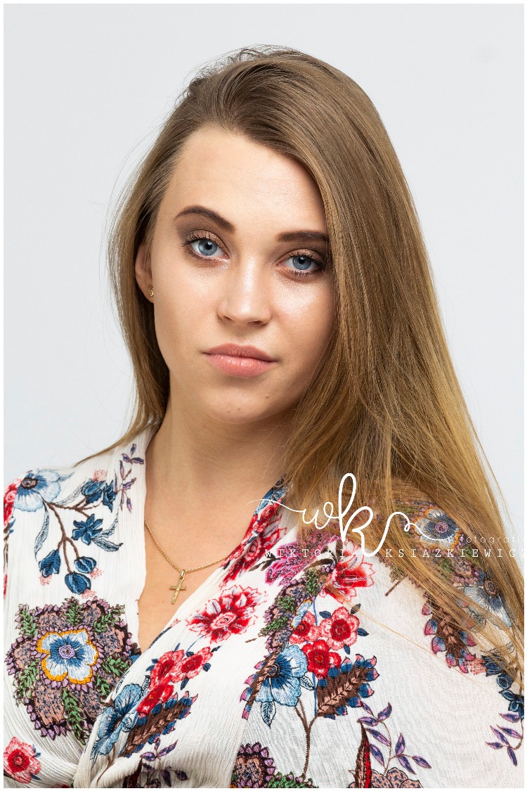 Portret kobiety z jasnymi włosami i delikatnym makijażem oczu, ubrana w bluzkę w kwiaty, z widocznym złotym łańcuszkiem z krzyżykiem, na jasnym tle.