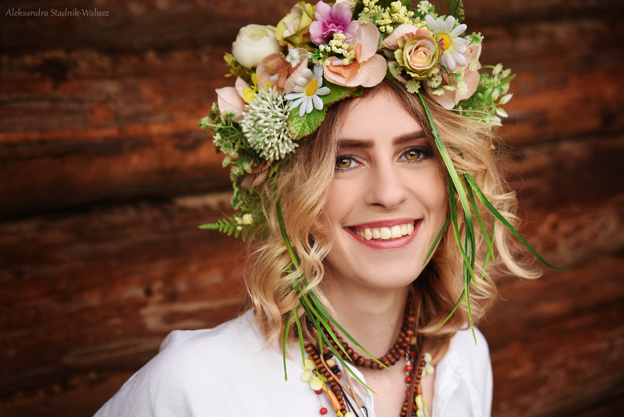Uśmiechnięta kobieta z zielonymi oczami w bogatym wianku z kwiatów i z delikatnym, naturalnym makijażem na tle drewnianej ściany.