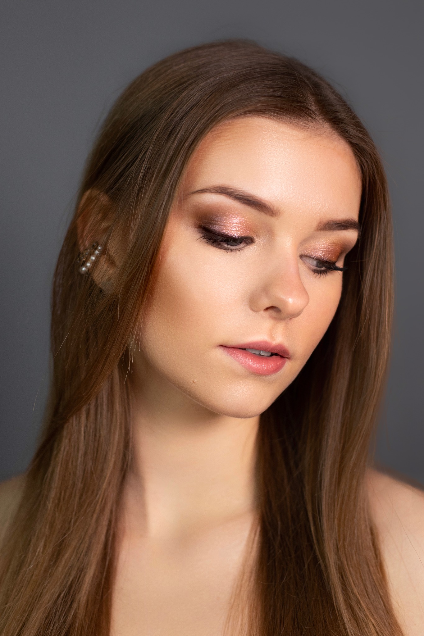 Portret młodej kobiety z profesjonalnym, błyszczącym makijażem oczu w odcieniach brązu i różu, delikatnie podkreślone usta, długie proste włosy w odcieniu miodowego brązu, subtelne kolczyki z pereł.