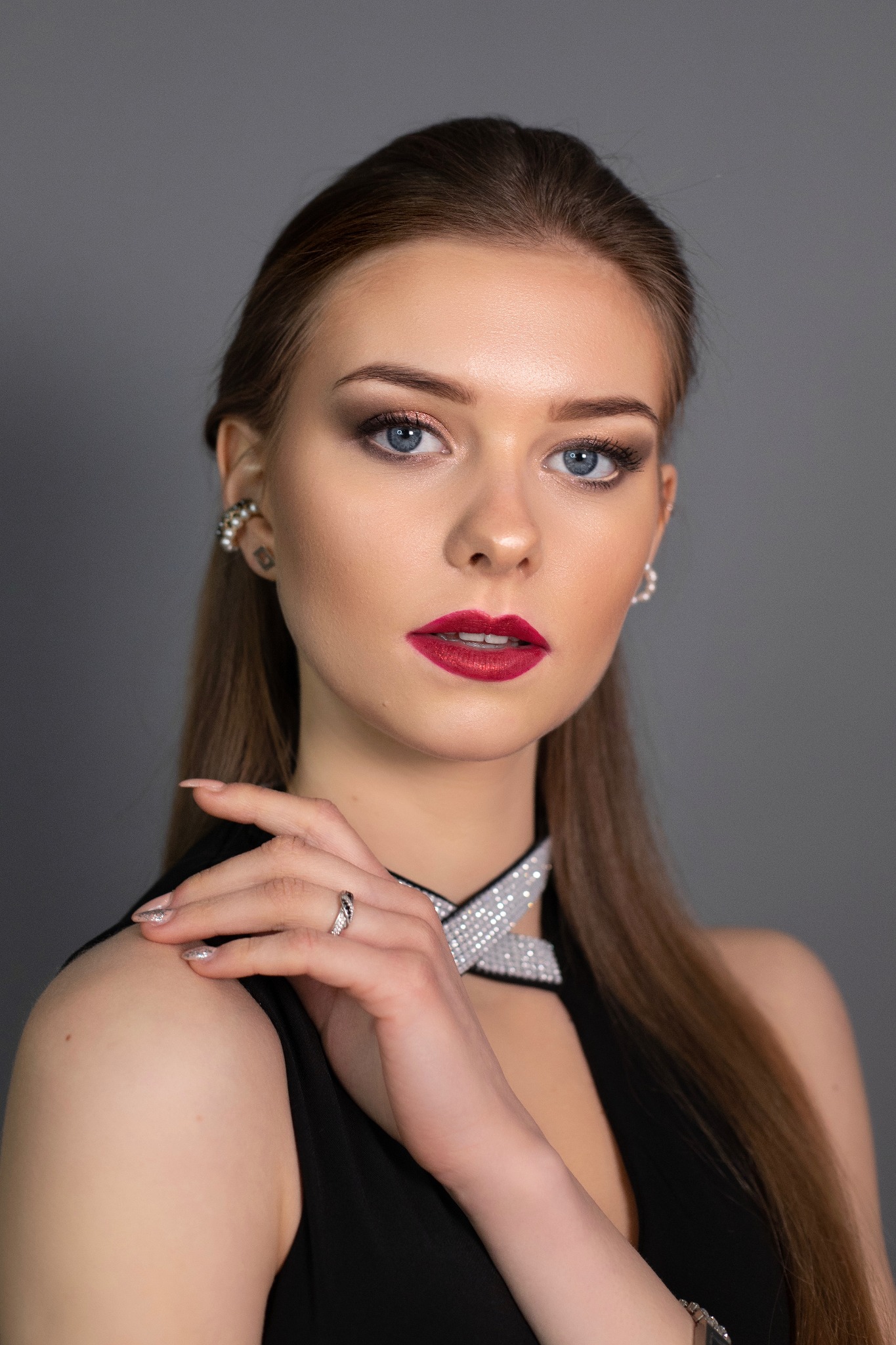 Portret kobiety z intensywnym makijażem oczu i ust, srebrną biżuterią i delikatnie pomalowanymi paznokciami na szarym tle.