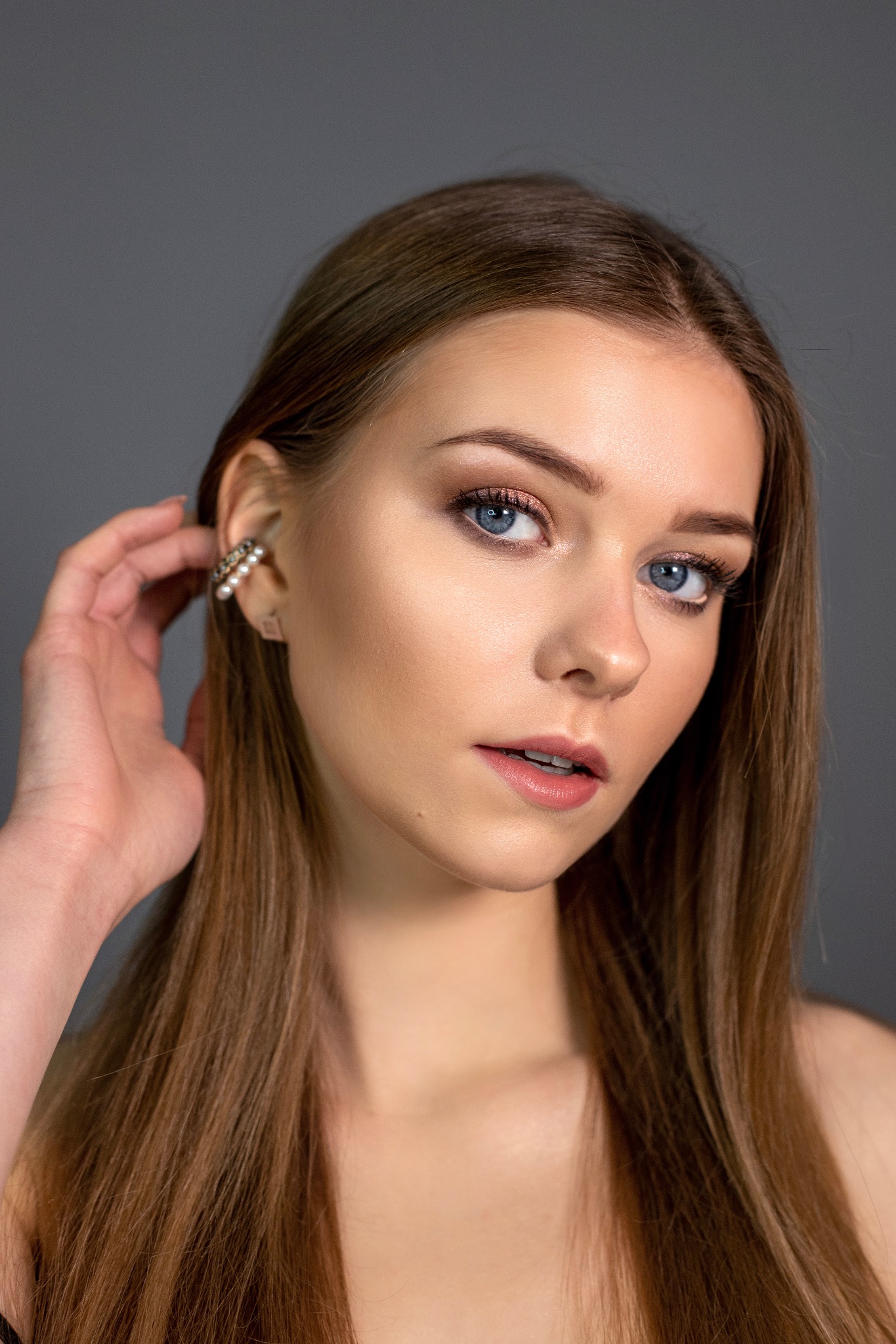 Portret kobiety z delikatnym makijażem oczu w odcieniach brązu i różu, podkreślonymi rzęsami, oraz subtelnym błyszczykiem na ustach. Ucho zdobi kolczyk typu cuff z perłami, a dłoń delikatnie dotyka...