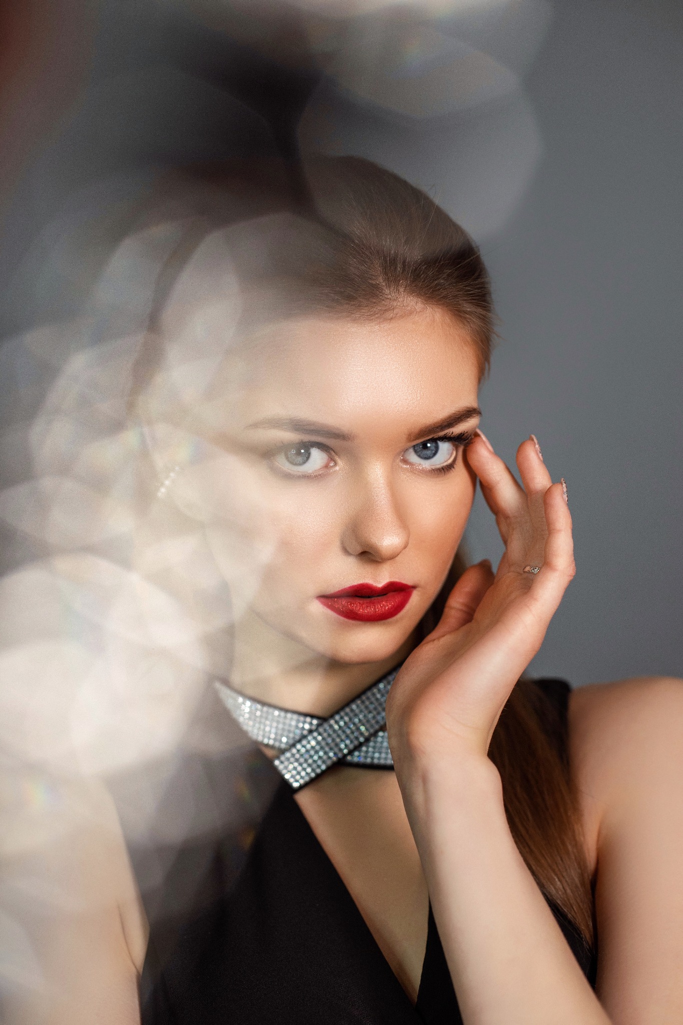 Portret kobiety z intensywnym czerwonym makijażem ust, subtelnym makijażem oczu, chokerem z kryształkami i artystycznym efektem rozmycia w tle.