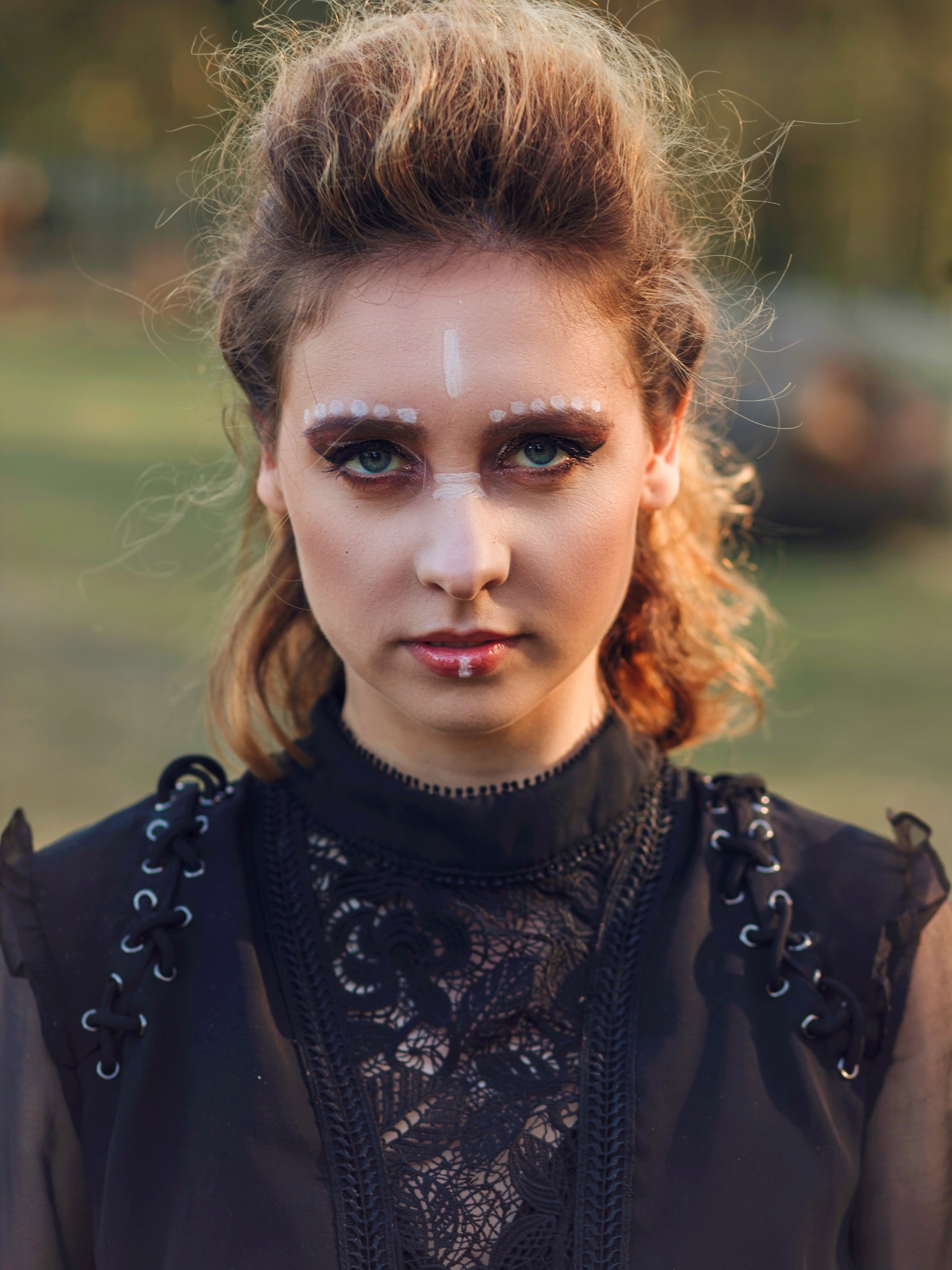 Portret kobiety z artystycznym, białym zdobieniem twarzy: linia na czole, kropki nad brwiami i kreska na dolnej wardze; ciemny makijaż oczu i upięte włosy w stylu vintage, na tle zieleni.