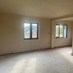 Surowe wnętrze po nałożeniu tynku maszynowego, widoczne okna z brązowymi ramami, wystające kable z sufitu i niedokończone wykończenie przy podłodze.