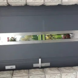 Szare drzwi wejściowe z poziomym, prostokątnym przeszkleniem ze stali nierdzewnej, przez które widać fragment ogrodu z kwiatami i ozdobnym ogrodzeniem.