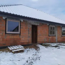 Surowy budynek w trakcie budowy, z zamontowanymi oknami z beżowymi ramami, przykryty śniegiem, widok z zewnątrz w zimowy dzień.