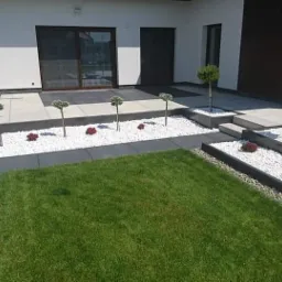 Nowoczesny taras z jasnych płyt, białych kamyków i ozdobnych drzewek, otoczony zielonym trawnikiem i tujami.