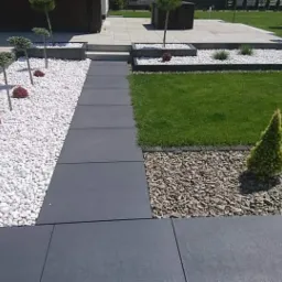 Nowoczesna aranżacja przestrzeni przed domem z ciemnoszarą ścieżką z płyt, białymi kamieniami, ozdobnymi drzewkami i trawnikiem