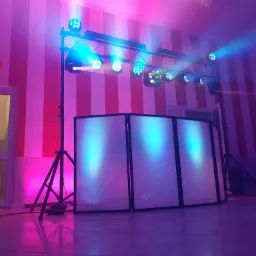 Scena z oświetleniem LED i panelem DJ-skim, podświetlona na różowo i niebiesko, tło w biało-czerwone pasy, po lewej stronie widoczne drzwi i dekoracja z balonów.