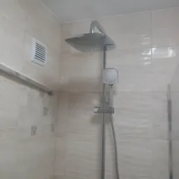 Zamontowany zestaw prysznicowy z deszczownicą i słuchawką na tle beżowych płytek w łazience z kratką wentylacyjną.